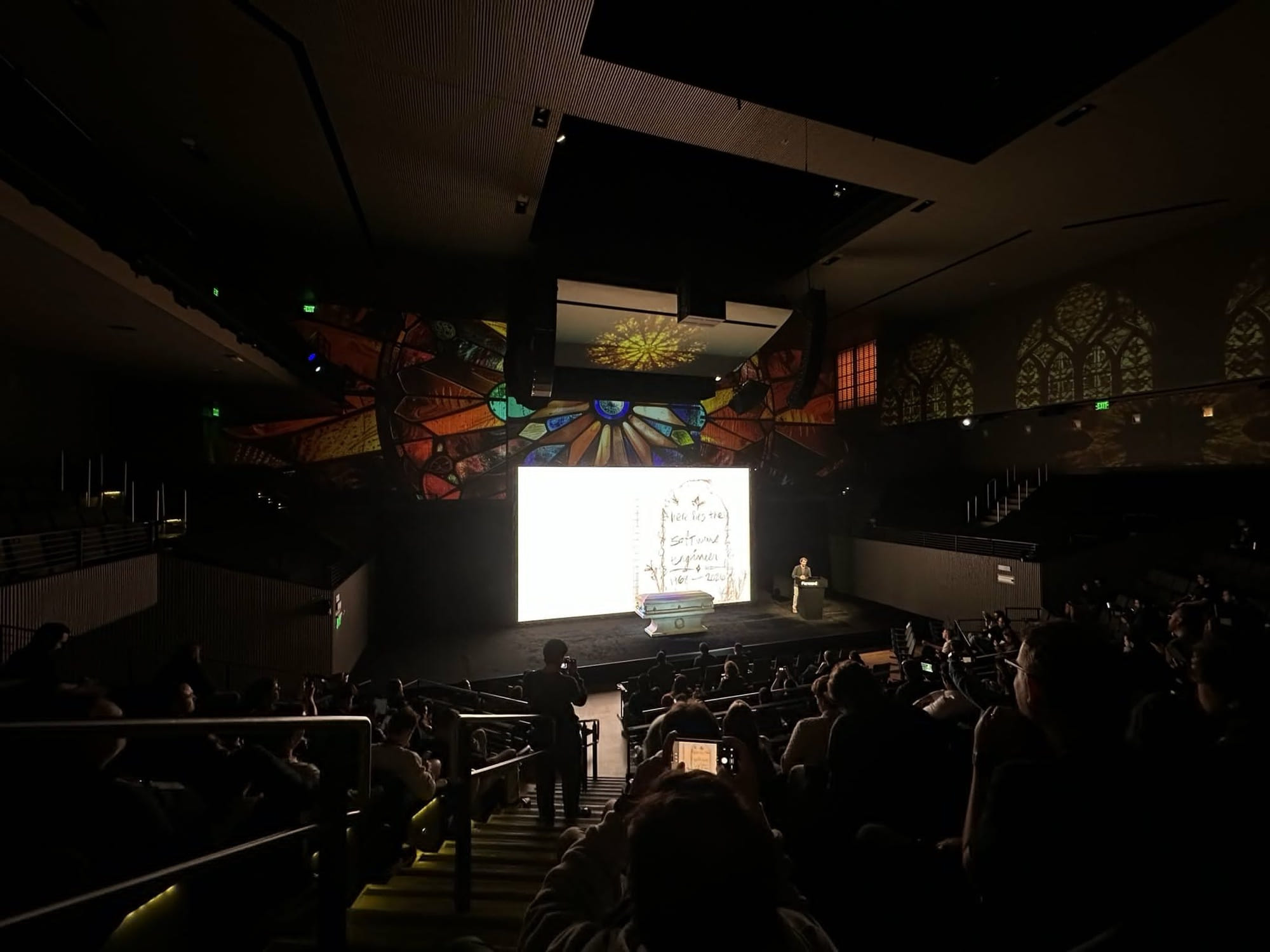 The Forward stage from the back of the room: the casket, the podium, and the pencil headstone on the big screen under projected stained glass